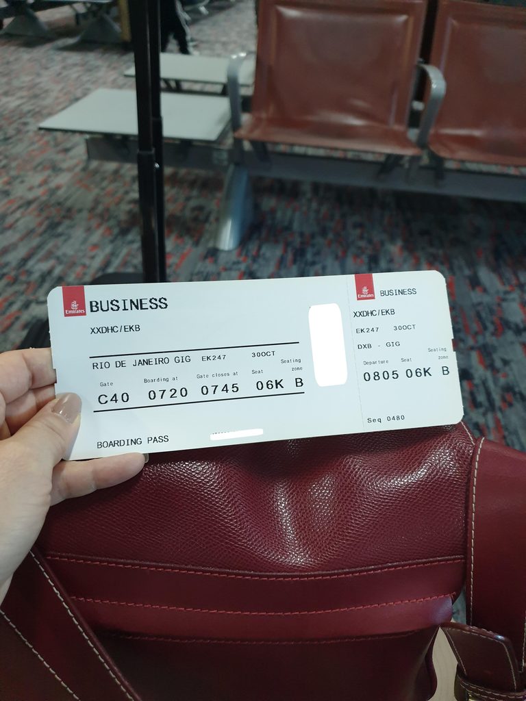 An Emirates boarding pass held in front of an airport departure board.